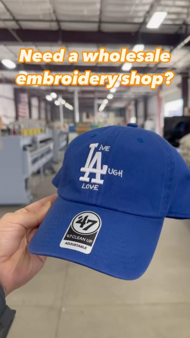 Hand holding a blue embroidered baseball cap with white text and graphics, in an industrial setting with the text overlay: "need a wholesale embroidery shop?.