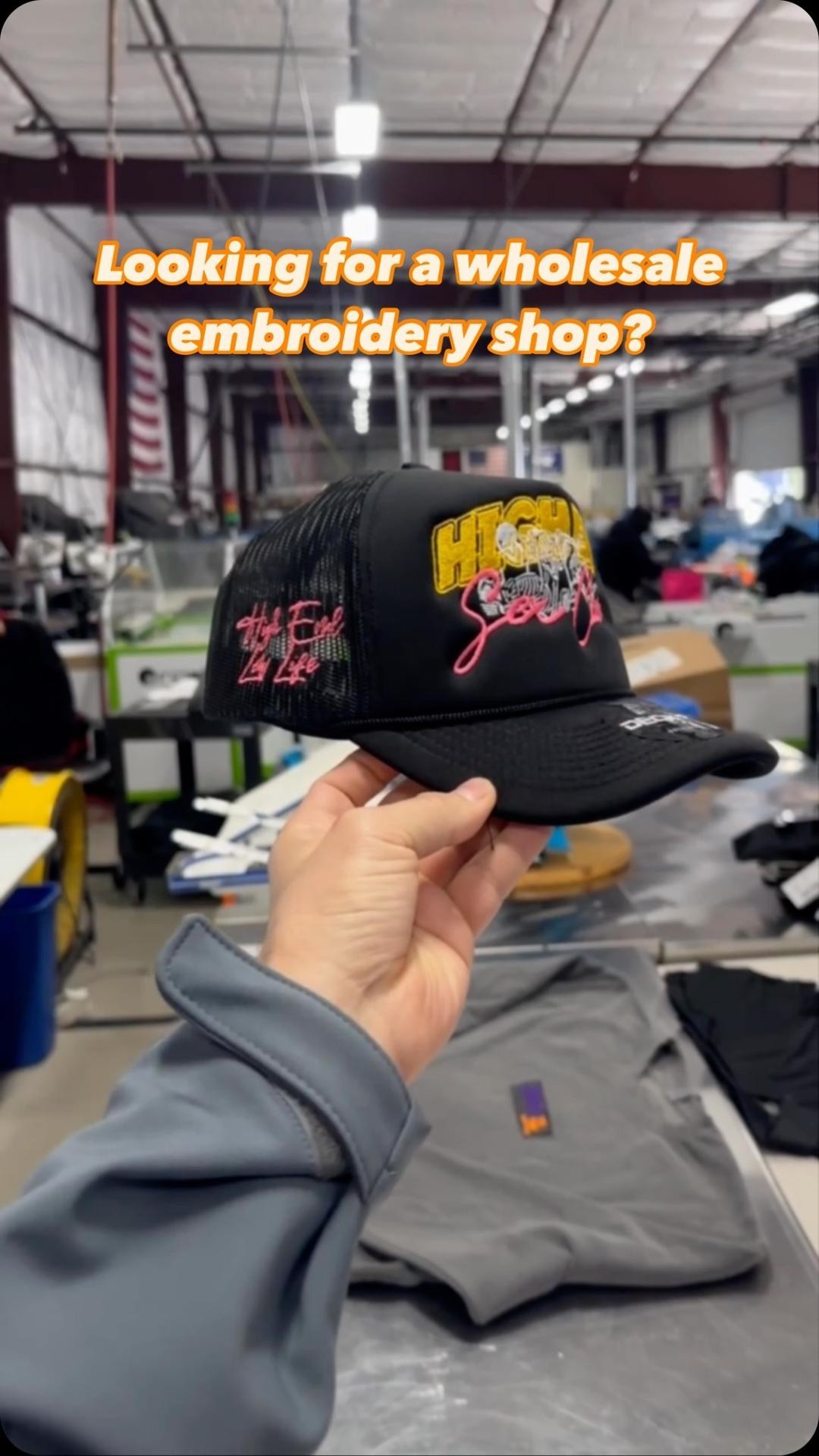 Hand holding a custom embroidered cap in a workshop environment with a promotional question about wholesale embroidery services.