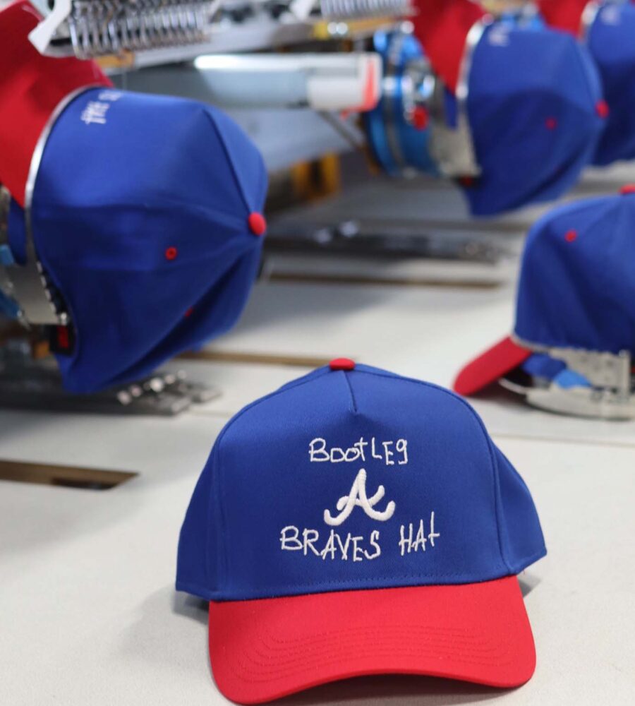 Embroidery machine in operation, producing blue and red baseball caps with white text and logo.