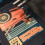 A graphic t-shirt with a vintage car design is being printed in a shop, focusing on the vivid colors and print details.