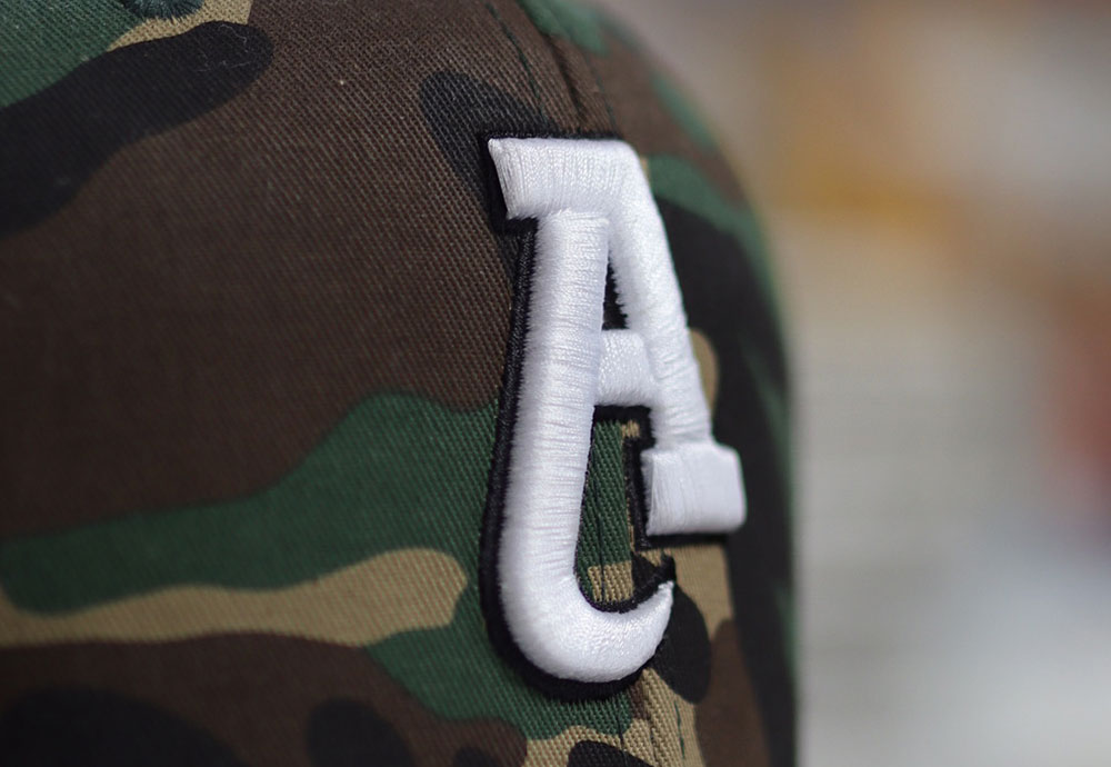 Close-up of a camouflage-patterned baseball cap with a large, white embroidered letter "A" on the side, showcasing our custom screen printing and embroidery services.