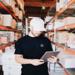A man in a black garment-decor t-shirt and white cap using a tablet in a warehouse with rows of shelves filled with boxes.