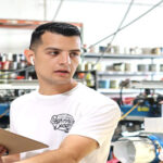 A man holding a clipboard in a workshop with shelves of garment-decor and mechanical parts in the background.