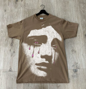 A brown t-shirt with a white graphic print of a stylized face featuring pink tear details, hanging on a hanger against a wooden floor background.
