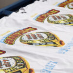 White t-shirts with "champion 2016" printed in bold colors on a conveyor belt in a factory.