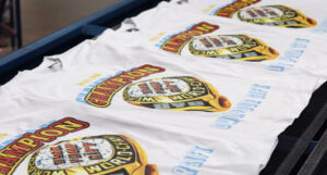 White t-shirts with "champion 2016" printed in bold colors on a conveyor belt in a factory.