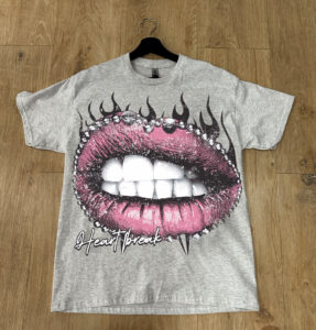 A gray t-shirt with a graphic print of pink lips and teeth, surrounded by black flames, and the text "heartbreak" displayed on a wooden floor.