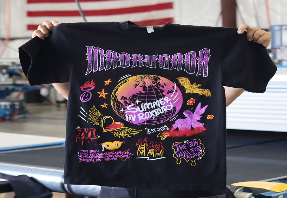 Person holding a black t-shirt with colorful garment-decor featuring the text "madrugada summer tour 2020" and various vibrant imagery including a globe, stars, and birds.