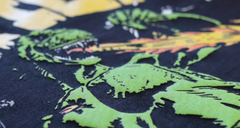 Close-up of a graphic t-shirt with a green and yellow dinosaur print, discovering types of print on a navy background.