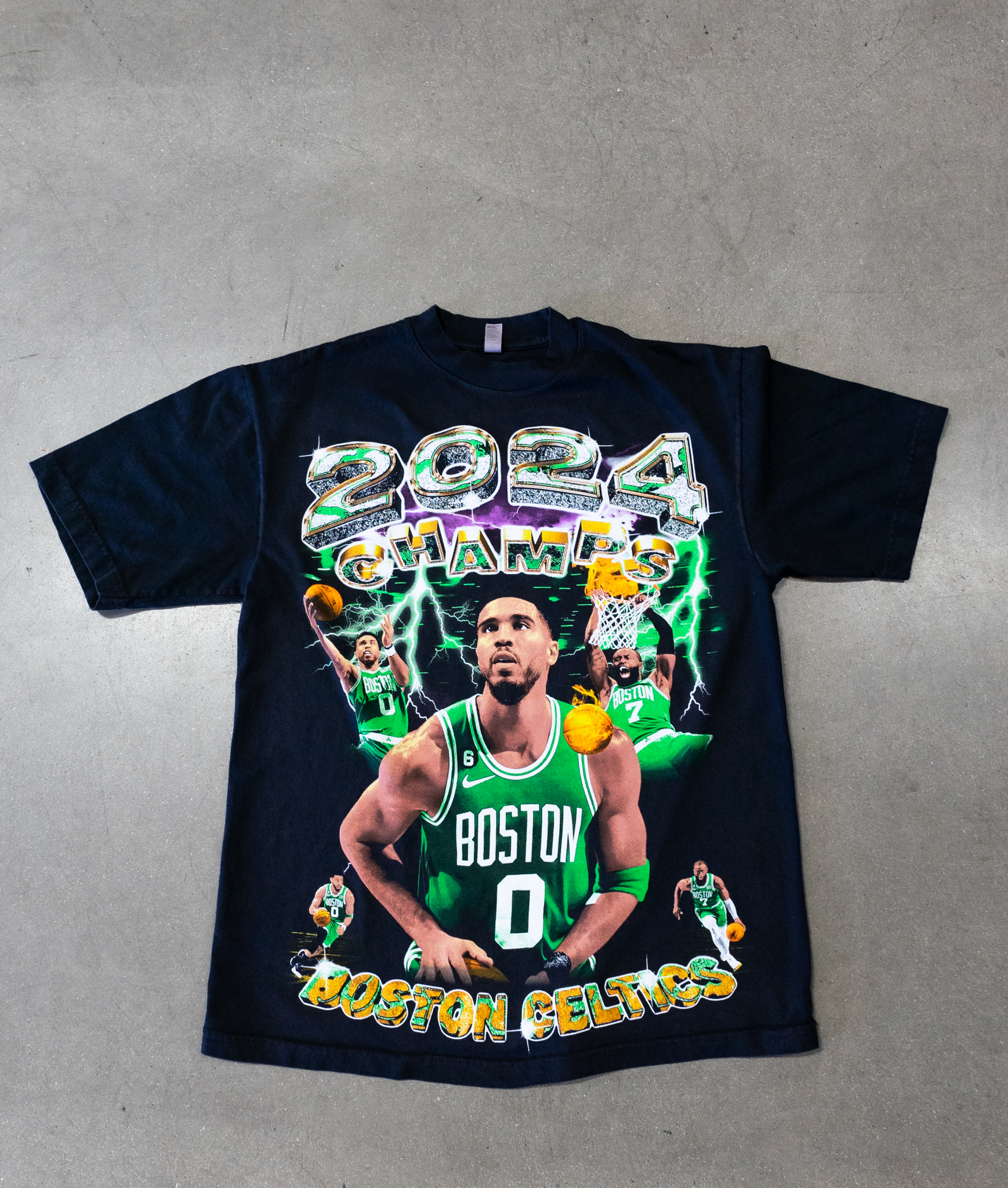 A black t-shirt with a screen-printed design featuring a basketball player in a green jersey, "2024 Champs" text above, and "Boston Celtics" text below. Various action poses of the player are also visible.