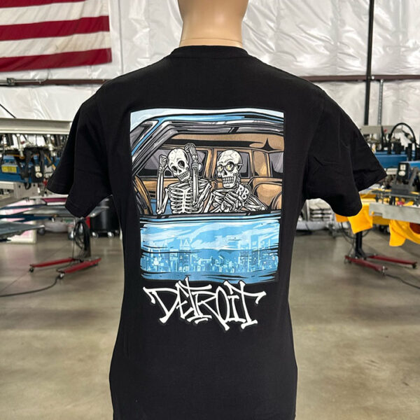 A mannequin displays a black 100 Digital Screen Printing- Garment-Dyed Heavyweight Shirt - Photo-Realistic Full-Color Printing - Hybrid Screen Print with Digital Color - 300 Resolution - Vivid Results with a graphic depicting two skeletons in a car, accompanied by the text "Detroit." An American flag is visible in the background.