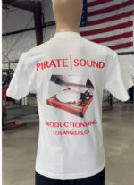 A mannequin wearing a 100 Digital Screen Printing- Garment-Dyed Heavyweight Shirt with "Pirate Sound Productions Inc. Los Angeles, CA" and an image of a turntable printed on the back. An American flag is visible in the background.