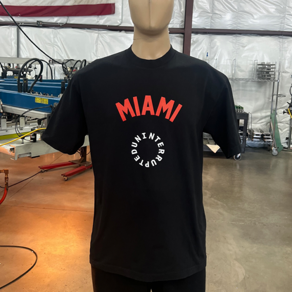 A mannequin displays one of the 100 Garment-Dyed Heavyweight Shirts - Text-Based Artwork with Vector Designs - Maximum of 2 Ink Colors - Soft Hand Screen Printing, featuring a black t-shirt with "MIAMI" in red text above and "INTERCEPTED" in white text below. The background includes an American flag and industrial equipment.