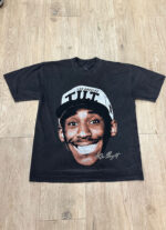A black t-shirt featuring a large, smiling face of a person wearing a hat with the text "Los Angeles" and unreadable text below. It lies flat on a wooden floor. 100 Custom Streetwear T-Shirts - Heavyweight Garment-Dyed Shirt With Screen Printing.