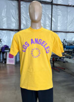 A mannequin is dressed in a 100 Garment-Dyed Heavyweight Shirt with Text-Based Artwork with Vector Designs - Maximum of 2 Ink Colors - Soft Hand Screen Printing in yellow, featuring the text "LOS ANGELES DOUNITE INT'L CREW" in purple. The mannequin is located in an industrial setting.