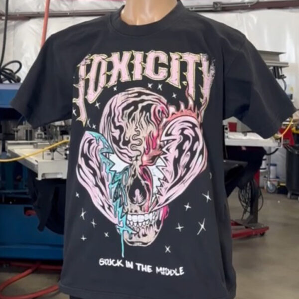 A mannequin displays a 100 Heavyweight Drop Shoulder Blanks - Streetwear - Maximum of 5 Ink Colors - Vibrant Loud Colors - Full Detail - Soft Hand Screen printing - Retail Ready with a colorful skull design and the word "TOXICITY" in bold letters, placed in an indoor setting with a U.S. flag partially visible in the background.