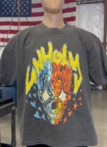 Mannequin wearing a 100 Heavyweight Drop Shoulder Blank with a graphic showing a half-blue, half-flaming skull with the word "UNHOLY" in bold, yellow letters above and surrounded by hearts and stars. American flag in background.