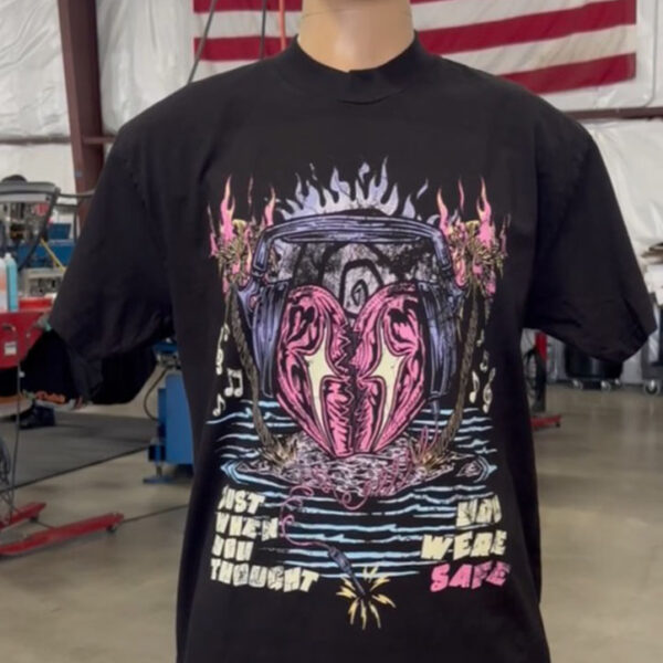 A mannequin dressed in a 100 Heavyweight Drop Shoulder Blanks - Streetwear - Maximum of 5 Ink Colors - Vibrant Loud Colors - Full Detail - Soft Hand Screen printing - Retail Ready featuring a vibrant, abstract design with text that reads "JUST WHEN YOU THOUGHT YOU WERE SAFE" displayed in an industrial setting with an American flag in the background.