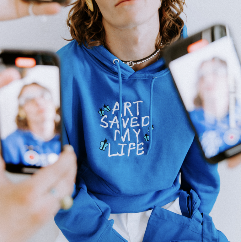 A person wearing Custom Embroidered Blue Lane Seven Apparel LS14001 Premium Pullover Hoodie with a design that reads "Art Saved My Life" on the front view - Garment Decor A person wearing Custom Embroidered Blue Lane Seven Apparel LS14001 Premium Pullover Hoodie with a design that reads "Art Saved My Life" on the front view