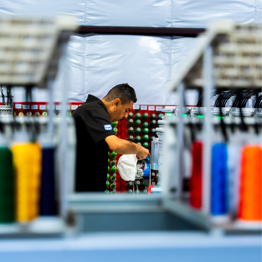 A man doing a quality check on a custom embroidered Arrowhead Evaluation Services, Inc. logo on a white Valucap VC600 Chino Cap embroidered by Garment Decor using a Barudan machine